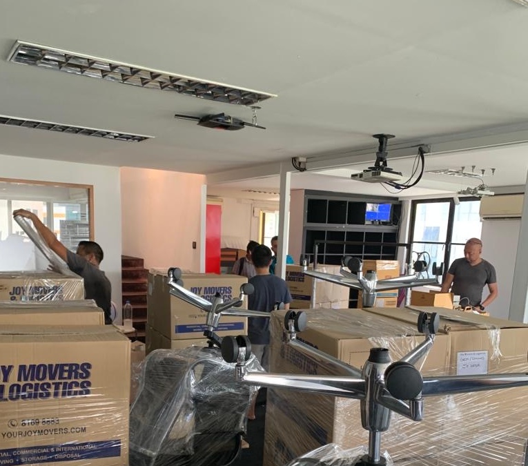 Several movers are packing and organizing cardboard boxes and office furniture in a well-lit room.